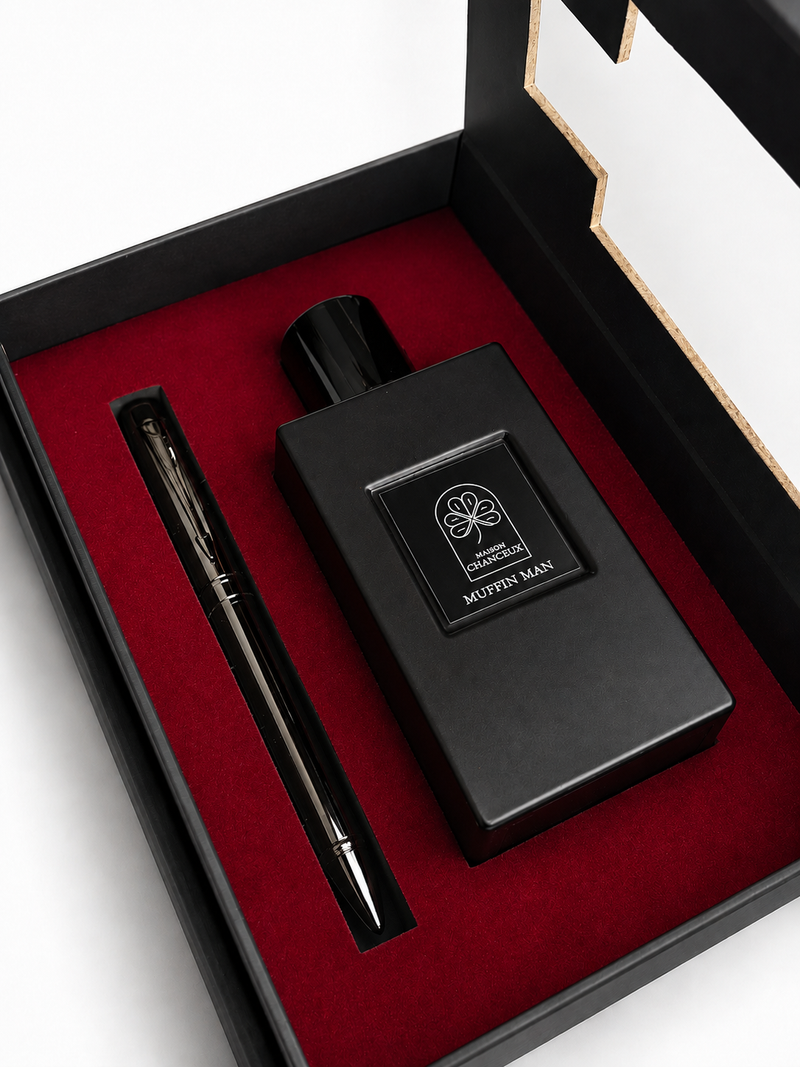A luxury gift set featuring a matte black rectangular perfume bottle with a silver logo and a matching black pen, presented in an open black box lined with deep red velvet.
